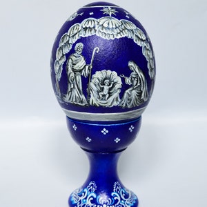 May include: A blue and white hand-painted wooden egg depicting the nativity scene. The egg is on a blue stand with white floral designs.