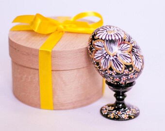 Op de afbeelding: Een handbeschilderd houten ei met een zwart, wit en gouden bloemenontwerp staat op een zwarte houten standaard. Het ei wordt gepresenteerd naast een houten geschenkdoos met een geel lint.