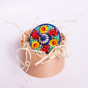 Puede incluir: Una peonza de madera pintada de azul y blanco con dise&ntilde;os florales rojos y amarillos, sentada en una caja marr&oacute;n llena de papel triturado.