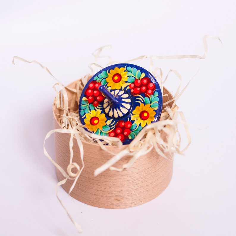 Puede incluir: Una peonza de madera pintada de azul y blanco con dise&ntilde;os florales rojos y amarillos, sentada en una caja marr&oacute;n llena de papel triturado.