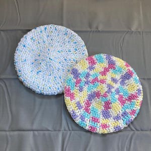 May include: Two round, crocheted items. One is white with blue and yellow flecks. The other is a colorful spiral of pink, yellow, blue, and purple. Both are on a gray surface.