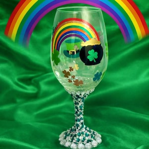 May include: A clear wine glass decorated with a rainbow, a leprechaun's hat, a pot of gold, and shamrocks. The stem is embellished with green and silver beads, and the base is adorned with white gems. A rainbow arches above the glass.