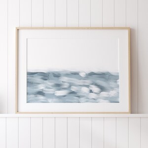 May include: A framed abstract painting of a blue and white seascape. The painting is in a light wood frame and is hanging on a white wall.