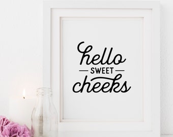 May include: White framed print with the text "hello sweet cheeks" in black script font. The print is on a white surface with a white candle, a clear glass bottle, and pink roses.