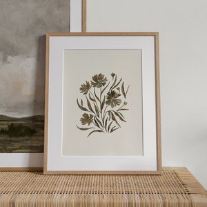 May include: A framed print of a watercolor painting of a bouquet of brown flowers on a white background. The print is in a light wood frame and is sitting on a woven wicker bench.