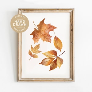 Peut inclure: Une impression aquarelle encadrée représentant quatre feuilles d'automne dans des tons orange, jaune et marron. Un badge circulaire dans le coin supérieur gauche contient le texte MADE BY A HAND DRAWN REAL ARTIST. Le cadre est en bois clair.