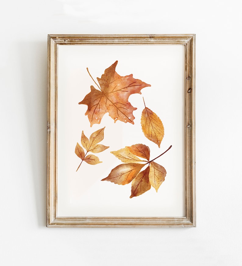 May include: A framed watercolor print featuring four autumn leaves in shades of orange, brown, and yellow. The leaves are arranged against a white background, enclosed in a rectangular wooden frame with a light brown, weathered finish.