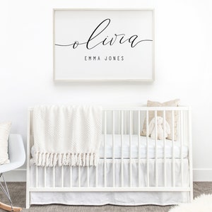 Op de afbeelding: Een wit babybedje met een wit dekentje en een beige kussen. Een ingelijst print met de naam Olivia en de tekst "Emma Jones" hangt aan de muur boven het bedje.