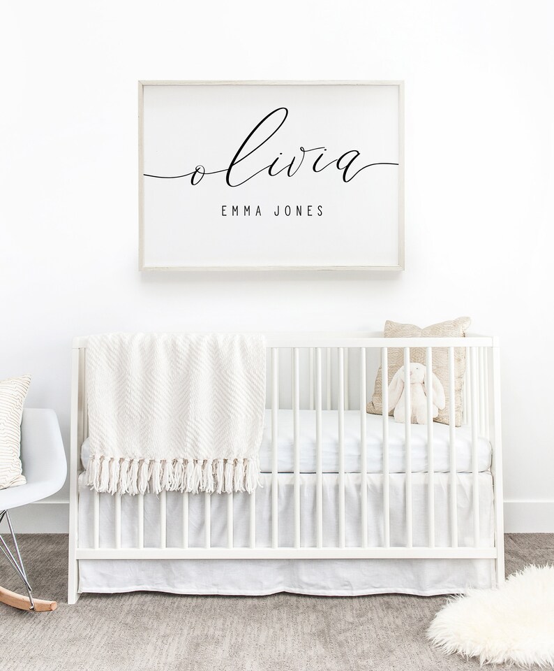 Op de afbeelding: Een wit babybedje met een wit dekentje en een beige kussen. Een ingelijst print met de naam Olivia en de tekst "Emma Jones" hangt aan de muur boven het bedje.