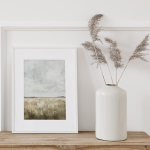 May include: A white framed picture with a painting of a field with a cloudy sky. The picture is sitting on a wooden table next to a white ceramic vase with dried grasses.