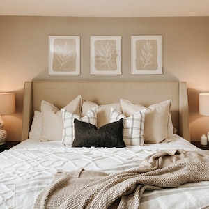 May include: A beige upholstered bed with a white duvet and a brown throw blanket. The headboard has three framed prints with a botanical design. The bed is styled with a variety of pillows in white, black, and grey.