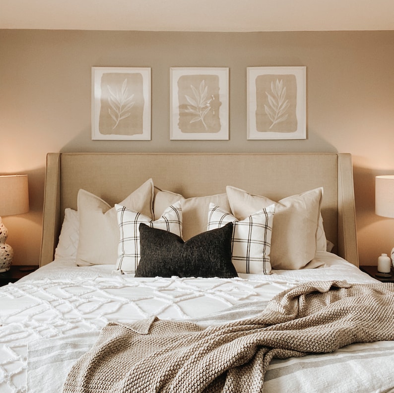 May include: A beige upholstered bed with a white duvet and a brown throw blanket. The headboard has three framed prints with a botanical design. The bed is styled with a variety of pillows in white, black, and grey.