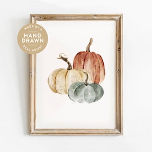 May include: A framed watercolor print featuring three pumpkins in shades of cream, orange, and muted green. A circular badge in the upper left corner contains the text MADE BY A HAND DRAWN REAL ARTIST. The frame is a light brown wood finish.