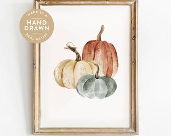 May include: A framed watercolor print featuring three pumpkins in shades of cream, orange, and muted green. A circular badge in the upper left corner contains the text MADE BY A HAND DRAWN REAL ARTIST. The frame is a light brown wood finish.