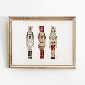 May include: Three watercolor nutcracker figurines in a wooden frame. The nutcrackers are painted in shades of red, white, and brown. The nutcracker on the left is wearing a white uniform with gold accents. The nutcracker in the middle is wearing a red uniform with gold accents. The nutcracker on the right is wearing a brown uniform with gold accents.