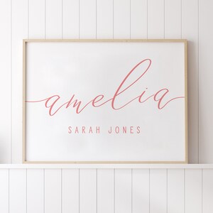 Op de afbeelding: Een ingelijst afdruk met de naam "amelia" geschreven in cursief in roze letters. Onder de naam staat de tekst "SARAH JONES" in een kleiner lettertype.