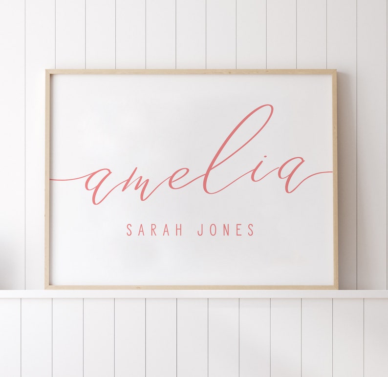 Op de afbeelding: Een ingelijst afdruk met de naam "amelia" geschreven in cursief in roze letters. Onder de naam staat de tekst "SARAH JONES" in een kleiner lettertype.