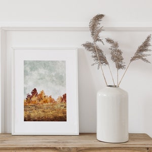 May include: A white frame containing a watercolour print of a woodland scene. The print depicts a pale blue sky with white clouds and a line of trees in shades of red, orange, and yellow. The frame is placed on a wooden surface alongside a white vase filled with dried grasses.