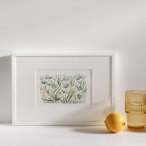 May include: A white framed print of a watercolor painting of blue flowers with green stems and leaves. The print is on a white background and is sitting on a white surface.