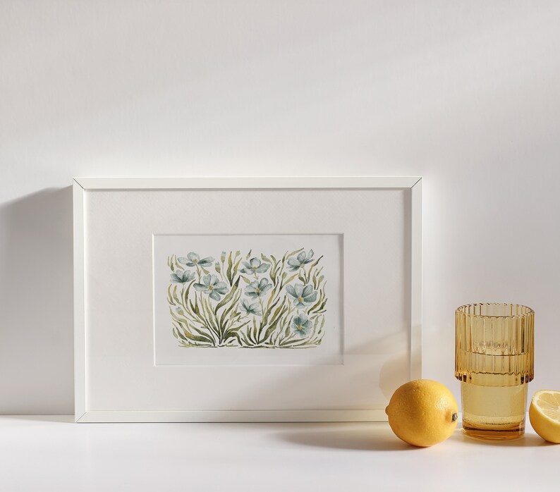 May include: A white framed print of a watercolor painting of blue flowers with green stems and leaves. The print is on a white background and is sitting on a white surface.