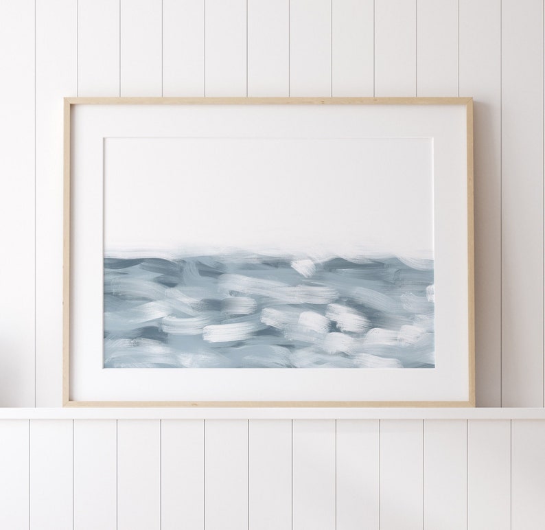May include: A framed abstract painting of a blue and white seascape. The painting is in a light wood frame and is hanging on a white wall.