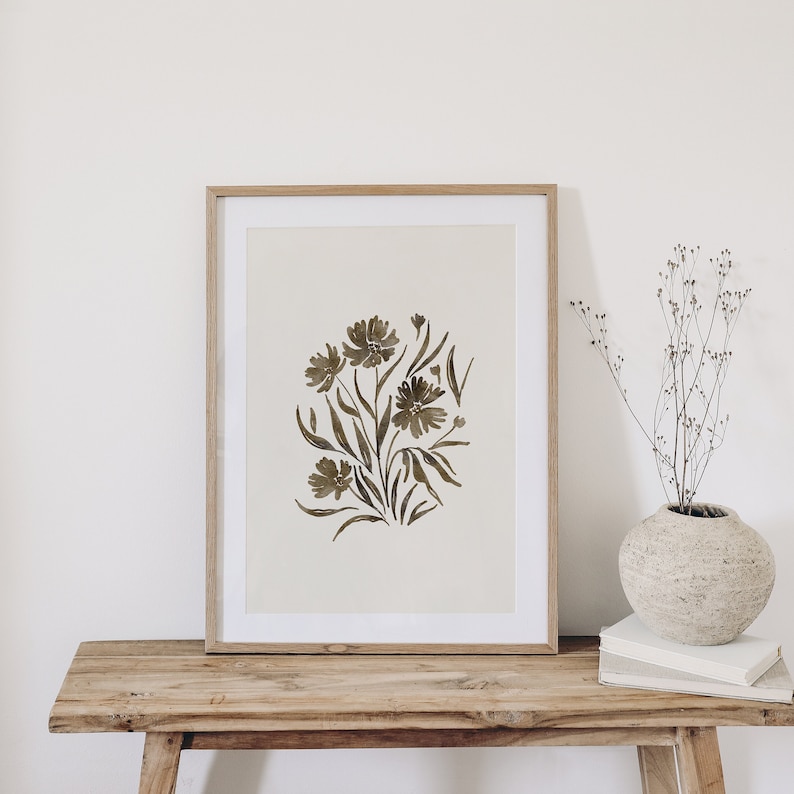 May include: A framed print of a watercolor painting of brown flowers with green stems and leaves on a white background. The print is in a light wood frame and is sitting on a wooden table.