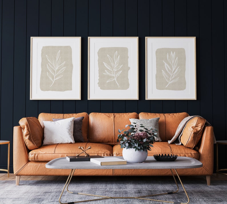 May include: Three framed prints with a beige background and white line drawings of leaves hang above a brown leather sofa with cushions. A small potted plant sits on a coffee table in front of the sofa.