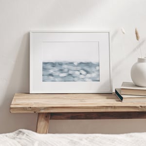 May include: A white framed print of a blue abstract painting depicting a seascape. The print is sitting on a wooden shelf with a vase of dried flowers and a stack of books.