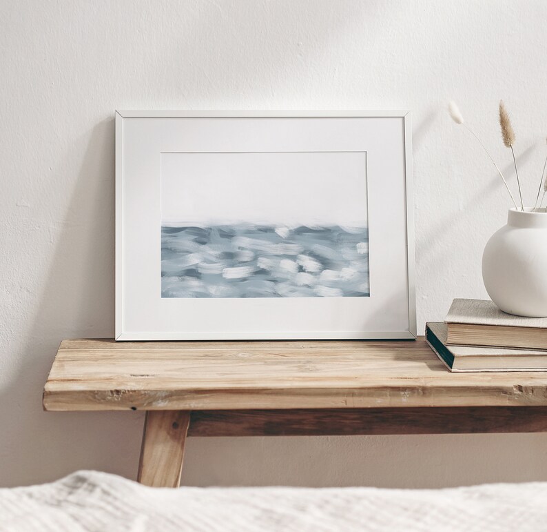 May include: A white framed print of a blue abstract painting depicting a seascape. The print is sitting on a wooden shelf with a vase of dried flowers and a stack of books.