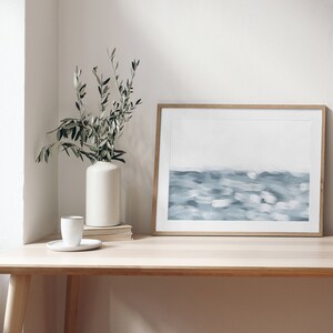 May include: A framed abstract painting of a blue and white seascape hangs on a light wood desk with a white vase and green plant in the corner.