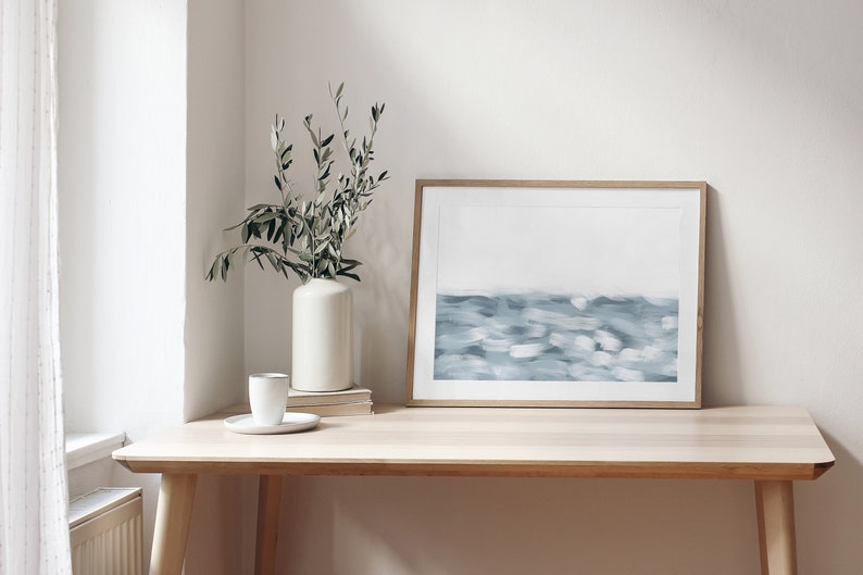 May include: A framed abstract painting of a blue and white seascape hangs on a light wood desk with a white vase and green plant in the corner.