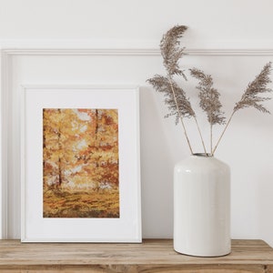 May include: A white frame with a print of a watercolor painting of a forest in autumn colors. The frame is sitting on a wooden table next to a white vase with dried grasses.