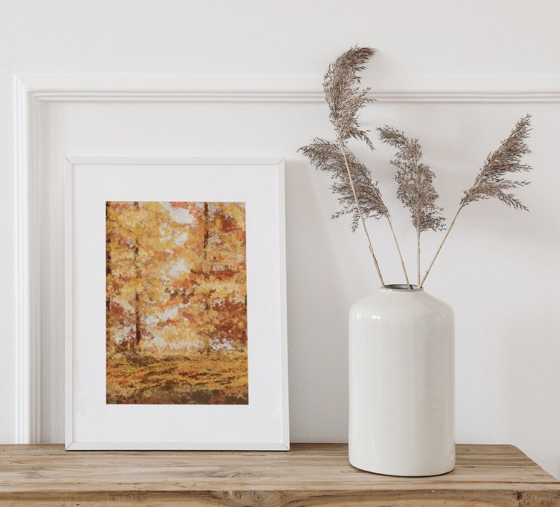 May include: A white frame with a print of a watercolor painting of a forest in autumn colors. The frame is sitting on a wooden table next to a white vase with dried grasses.
