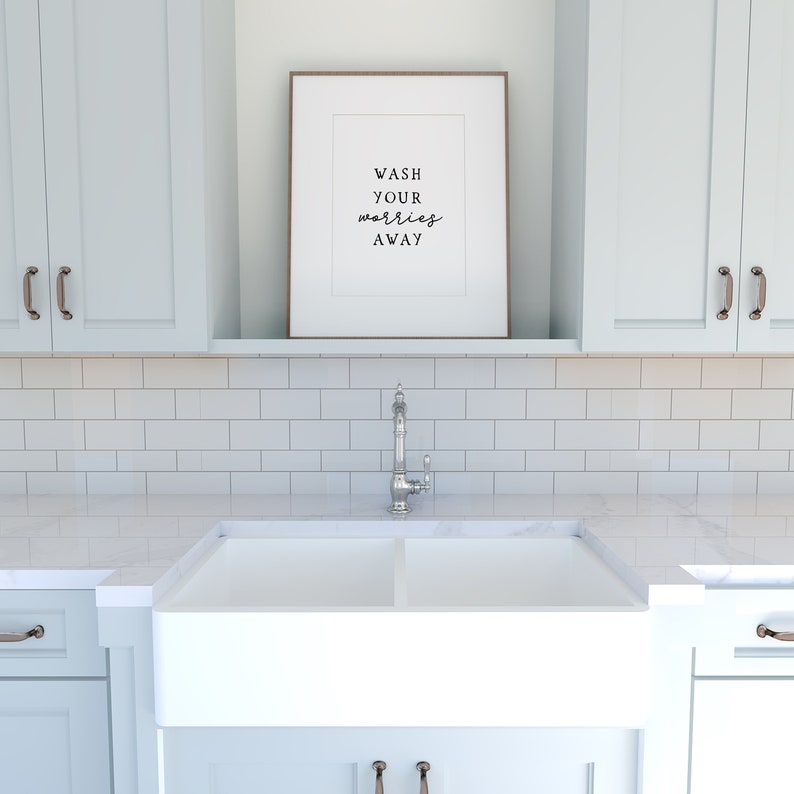 May include: A white farmhouse style kitchen sink with a chrome faucet. The sink is set against a white subway tile backsplash. A framed print with the text "Wash your worries away" hangs above the sink.