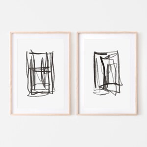 May include: Two framed black and white abstract art prints with a minimalist style. Each print features a square shape with black lines and strokes.