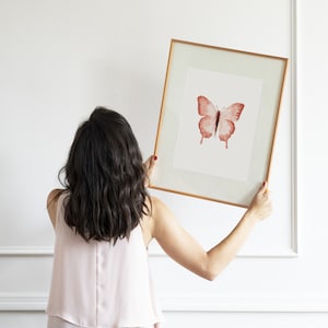 May include: A person hangs a framed print of a red butterfly on a white wall. The butterfly is centered in the frame and has a white background.
