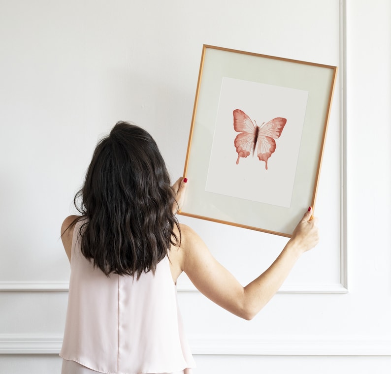 May include: A person hangs a framed print of a red butterfly on a white wall. The butterfly is centered in the frame and has a white background.