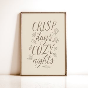 Op de afbeelding: Een ingelijst print met de tekst "CRISP days COZY nights" in bruine cursieve letters. De tekst wordt omringd door kleine blaadjesillustraties.