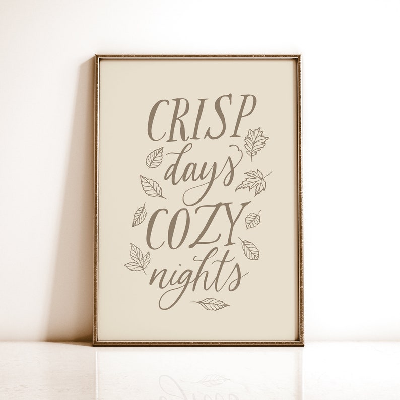 May include: A framed print with a beige background and brown text that reads 'CRISP days COZY nights' with small leaf drawings surrounding the text.