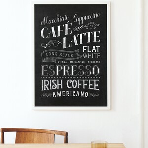 May include: Black and white chalkboard style sign with a white frame listing different types of coffee drinks, including Macchiato, Cappuccino, Cafe Latte, Flat White, Long Black, Vienna, Mochaccino, Affogato, Espresso, Irish Coffee, and Americano. The sign is on a wooden table with a glass of water and a chair.