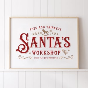 Puede incluir: Un cartel blanco con marco de madera marrón con un diseño rojo y dorado. El cartel dice "Juguetes y baratijas El taller de Santa Claus Calle del Caramelo, Polo Norte".