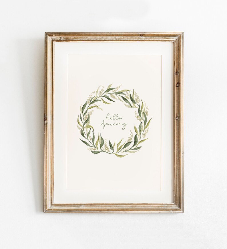 May include: A framed print with a watercolor illustration of a green leafy wreath with white flowers. The wreath surrounds the text "hello spring".