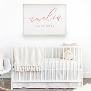 Op de afbeelding: Een wit babybedje met een wit dekentje en een wit knuffelkonijn. Een ingelijst print met de naam "amelia" in roze schrift en de tekst "SARAH JONES" in kleinere roze schrift.
