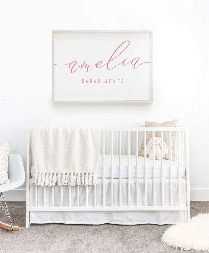 Op de afbeelding: Een wit babybedje met een wit dekentje en een wit knuffelkonijn. Een ingelijst print met de naam "amelia" in roze schrift en de tekst "SARAH JONES" in kleinere roze schrift.