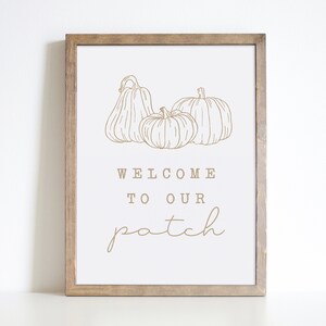 May include: A white sign with a brown wooden frame, featuring a line drawing of three pumpkins and the text "Welcome to our patch".
