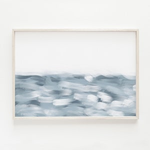 May include: A framed abstract painting of a blue ocean scene with white brushstrokes. The painting is in a white frame and is hanging on a white wall.