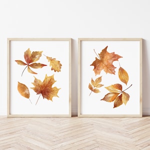 May include: Two framed wall art prints featuring watercolor illustrations of autumn leaves. Each print displays a collection of maple and oak leaves in shades of orange, yellow, and brown against a white background, housed in light wood frames.