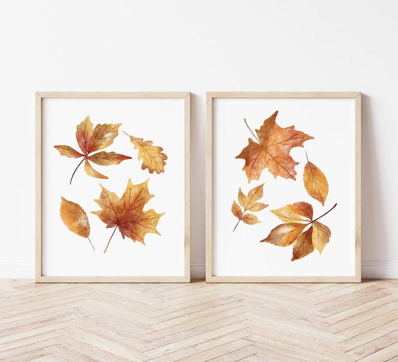 May include: Two framed wall art prints featuring watercolor illustrations of autumn leaves. Each print displays a collection of maple and oak leaves in shades of orange, yellow, and brown against a white background, housed in light wood frames.