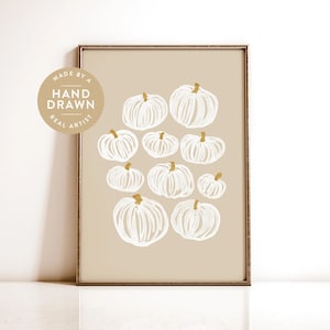 Può includere: Una stampa artistica da parete incorniciata raffigurante dieci zucche bianche disegnate a mano con steli dorati su uno sfondo beige a tinta unita. Un badge circolare nell'angolo in alto a sinistra contiene il testo MADE BY A HAND DRAWN REAL ARTIST.