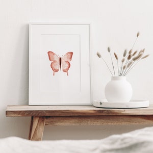 May include: A framed watercolor painting of a pink butterfly on a white background. The painting is displayed on a wooden shelf with a white vase of dried grasses.